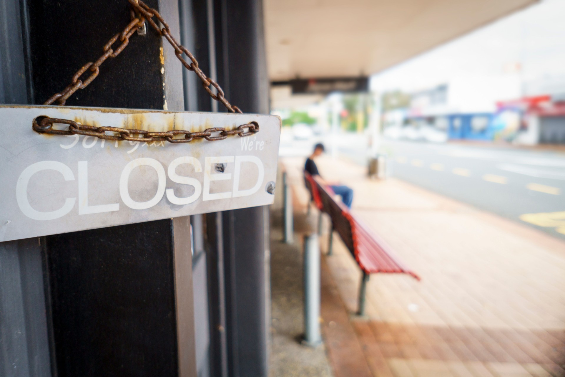 we are closed sign hanging on shop door. Unrecognizable person sitting on the bench outside.